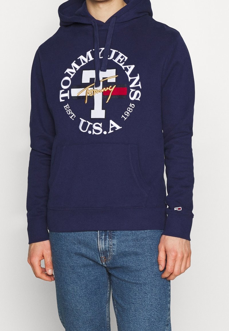 Personne portant un sweat à capuche bleu marine Tommy Jeans avec logo blanc et un jean, debout devant un fond uni clair.