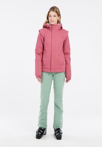 Rosa Skijacke mit hohem Kragen und verstellbarer Kapuze, getragen über hellgrünen Skihosen. Schwarze Skischuhe vervollständigen das Outfit.