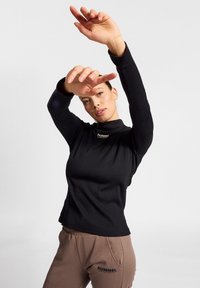 Zwarte langemouwtop met geribbelde textuur en hoge kraag, gecombineerd met bruine sweatpants met een logo. Armen omhoog geplaatst.