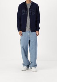 Sudadera con cremallera en color navy con el logo "GAP", camiseta gris debajo, combinada con jeans sueltos de color azul claro y zapatillas blancas. Tela suave, corte casual.