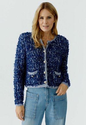 Femme aux cheveux blonds portant un cardigan bleu marine texturé boutonné et un jean bleu clair avec de grandes poches avant, sur un fond uni.