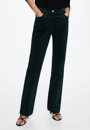 Pantalones rectos de pana verde oscuro, usados con sandalias negras de tiras y con las manos en los bolsillos, sobre fondo blanco.