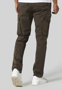 Mørkebrune cargo-bukser med flere sidelommer, tapered design og teksturert finish, sammen med hvite sneakers.