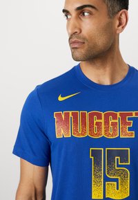 Zila kokvilnas T-kreklu ar drosu dzeltenu un sarkanu "NUGGETS" tekstu un numuru "15". Nike logs dzeltenā krāsā kreisajā plecā. Īsas piedurknes.