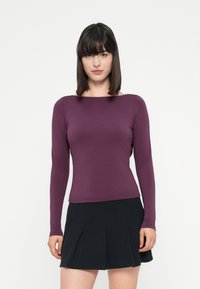 Donna con capelli lunghi e lisci di colore scuro, che indossa un top viola a maniche lunghe e una gonna nera plissettata, in piedi contro uno sfondo bianco.