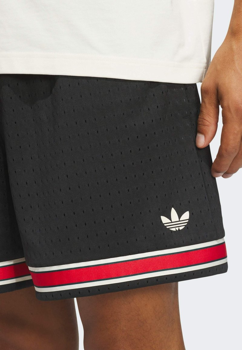 Schwarze Sportshorts mit kleinen Perforationen, rot-weißen Streifen am Saum und weißem Adidas-Trefoil-Logo an der Seite, getragen mit weißem Shirt.