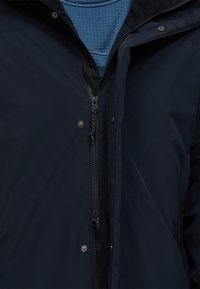 Marineblauwe jas met een volledige rits en drukknoopsluiting, met een gladde textuur en een hoge kraag. Draagt over een blauw patroonhemd.