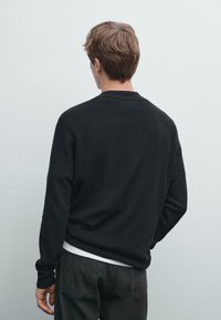 Zwarte gebreide trui met een ronde hals, lange mouwen en een geribbelde textuur. Gecombineerd met grijze jeans, gezien van de achterkant.