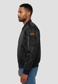 Zwarte bomberjack met een gladde afwerking, een ritsvak op de linkermouw en oranje tekstdetail op de schouder. Draagt blauwe spijkerbroek.