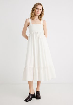 Γυναίκα με ξανθά μαλλιά μήκους μέχρι τους ώμους που φοράει λευκό μίντι φόρεμα χωρίς μανίκια και μαύρα μυτερά mules με τακούνι, στέκεται μπροστά σε απλό φόντο.