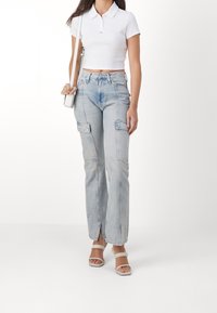 Kvinna i en vit kortärmad polo, ljusblå högmidjade cargobyxor med framsprund, vita klacksandaler och bärande en liten vit axelväska.