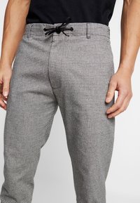 Pantalon à motif pied-de-poule gris avec une taille élastique et un cordon de serrage. Comprend des poches latérales et une coupe ajustée.