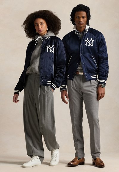 Két modell sötétkék New York Yankees dzsekit visel szürke nadrággal; az egyik modell fehér tornacipőt, a másik barna mokaszint hord.