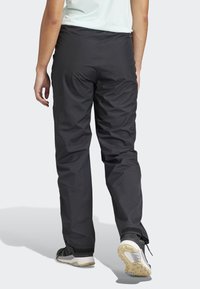 Pantalons étanches noirs avec une coupe décontractée, présentés avec une taille élastique et des poignets de cheville en Velcro, assortis à des chaussures de sport grises.