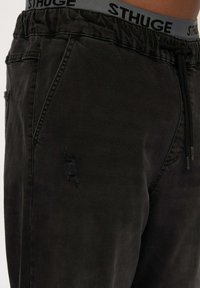 Pantalones de chándal negros hechos de una tela suave, con cintura ajustable por cordón, bolsillos laterales y un parche desgastado en el muslo.