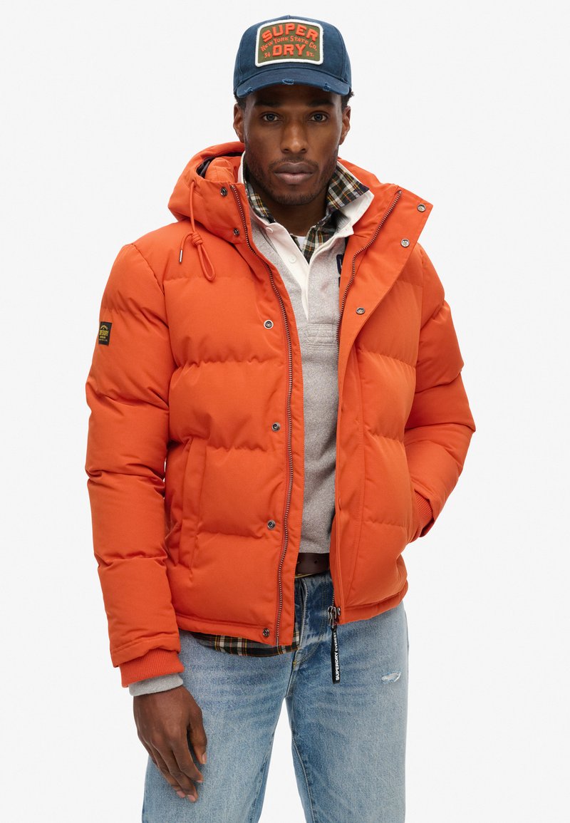Orange pufferjakke med hætte, der har hævede paneler, frontlynlås og trykknapper. Båret over en grå sweater og en rutet skjorte.