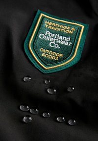 Czarna tkanina z zieloną i złotą haftowaną naszywką, na której widnieje napis "Dziedzictwo i Tradycja, Portland Outerwear Co." z kroplami wody na powierzchni.