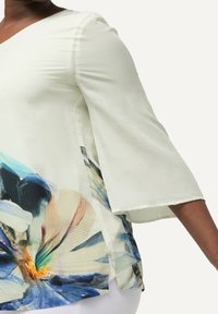 Blusa verde chiaro con maniche a campana, caratterizzata da un vivace motivo floreale in blu, arancione e bianco sui lati, realizzata in un tessuto liscio.