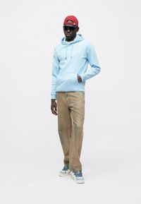 Gaiši zils džemperis ar kapuci un priekšpusi kabatu, bēša krāsas plašas bikses un gaiši zili sporta apavi. Izskatu papildina sarkans cepure un tumši saulesbrilles.