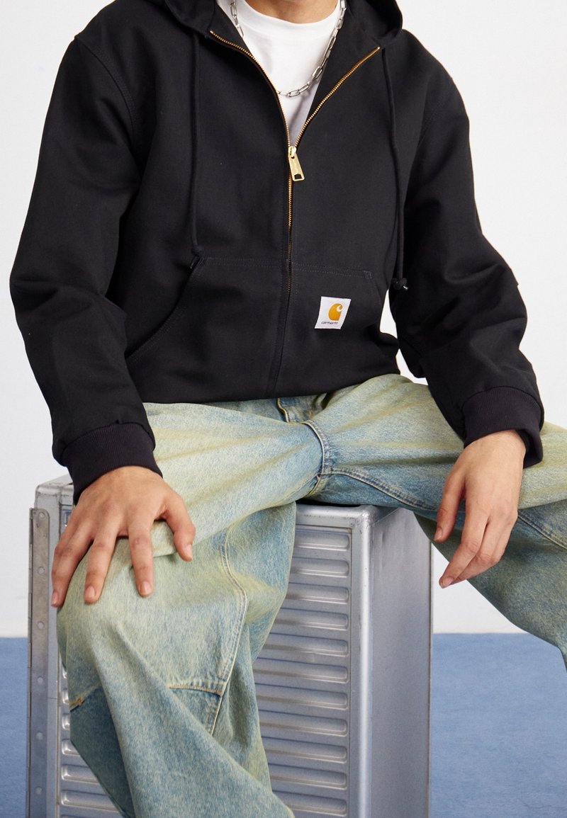 Schwarzer Kapuzenpullover mit Reißverschluss, Fronttaschen und einem Carhartt-Logo, kombiniert mit hellblauen, weiten Jeans. Strukturierter Stoff und lässige Passform.