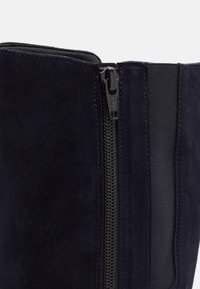 Botín de ante azul marino con cremallera lateral y panel elástico. Textura suave con acentos de costura a lo largo de las costuras.