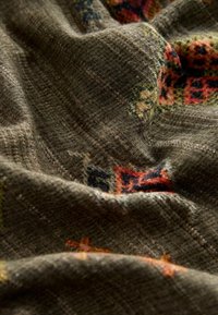 Zacht, gestructureerd olijfgroen stof met ingewikkelde meerkleurige geometrische en bloemenpatronen, getoond in close-up plooien en kreukels.