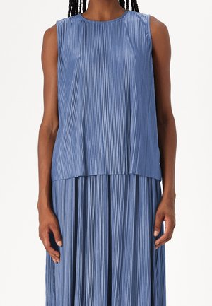 Femme portant une robe bleue plissée sans manches avec des lignes verticales et un col rond, montrée des épaules à la taille.