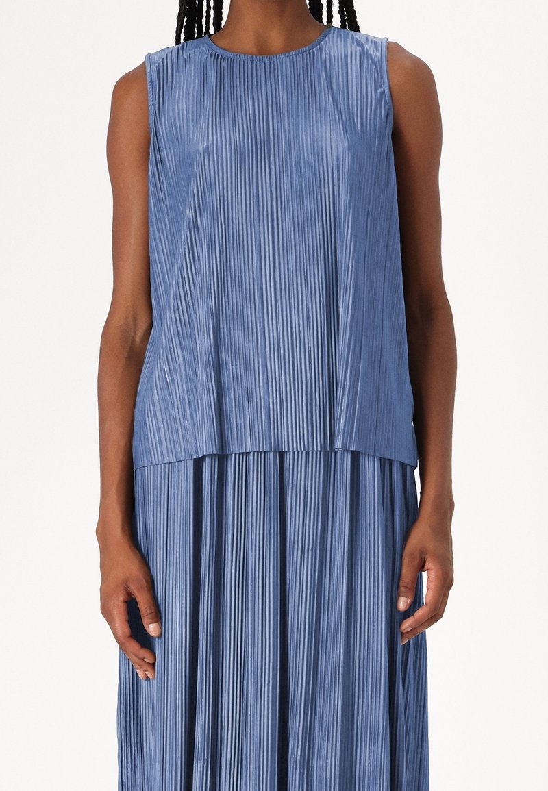 Femme portant une robe bleue plissée sans manches avec des lignes verticales et un col rond, montrée des épaules à la taille.