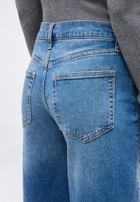 Κοντινό πλάνο από την πίσω όψη ενός ατόμου που φοράει μπλε τζιν και γκρι πλεκτό πουλόβερ με ριμπ λεπτομέρειες, εμφανίζοντας τις τσέπες και τις ραφές.