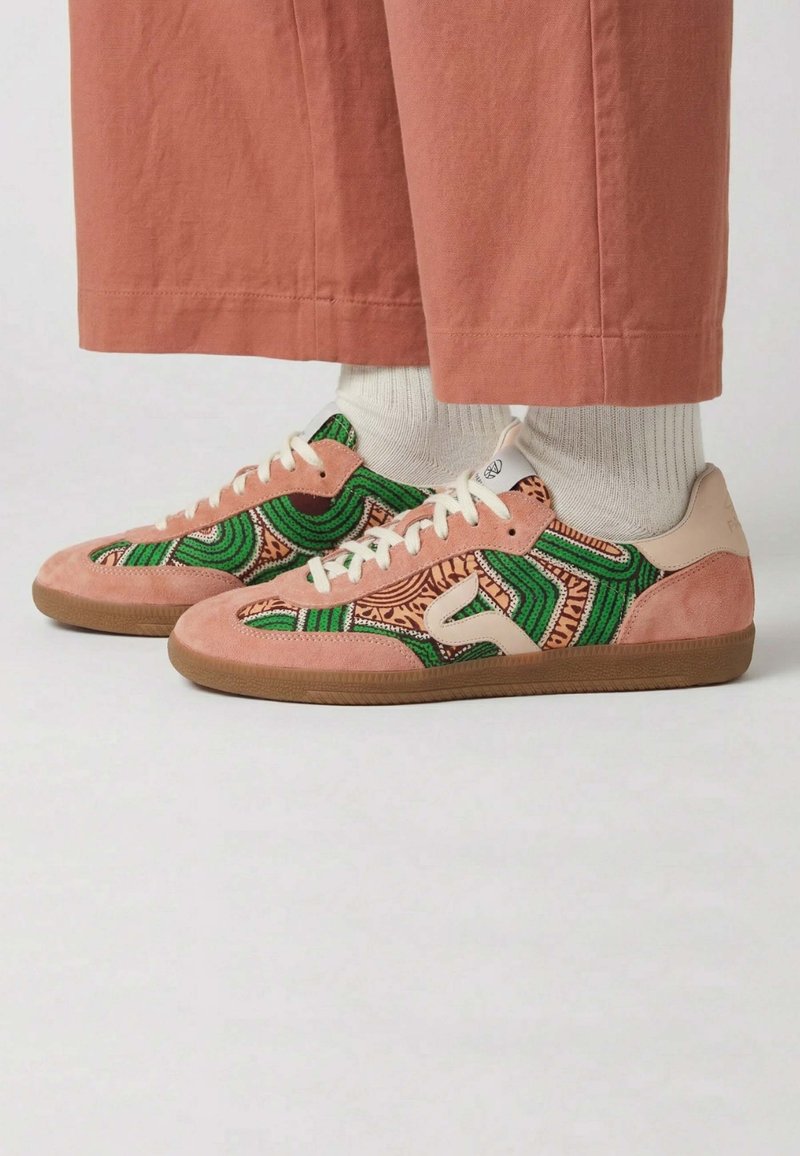 Pieds portant des baskets en daim et à motifs verts, roses et beige, avec des chaussettes blanches et un pantalon rose raccourci sur un fond clair uni.