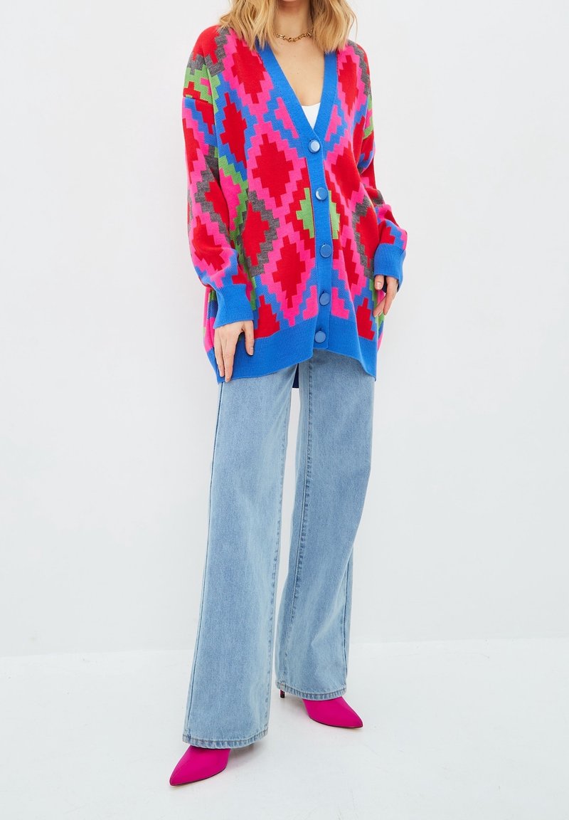 Kleurrijke gebreide cardigan met een ruitpatroon in roze, groen en blauw, voorzien van grote knopen en wijde mouwen, gedragen met lichtblauwe jeans.