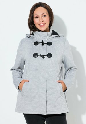 Žena se usmívá, má ruce v kapsách, nosí světle šedý kabát s kapucí a černými knoflíky a černé kalhoty, na bílém pozadí.