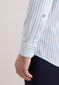 Weißes Hemd mit hellblauen vertikalen Streifen. Mit Knopfmanschetten und einem kleinen gestickten Motiv am Ärmel. Glatte Stofftextur.