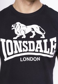 Fekete póló fehér oroszlán grafikával és vastag betűkkel írt "LONSDALE LONDON" felirattal. Klasszikus kerek nyakú dizájn, pamutból készült.