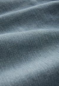 Denim tkanina u srednje plavoj nijansi, s glatkom teksturom i suptilnim dijagonalnim uzorcima tkanja te malo sjajnom završnom obradom.