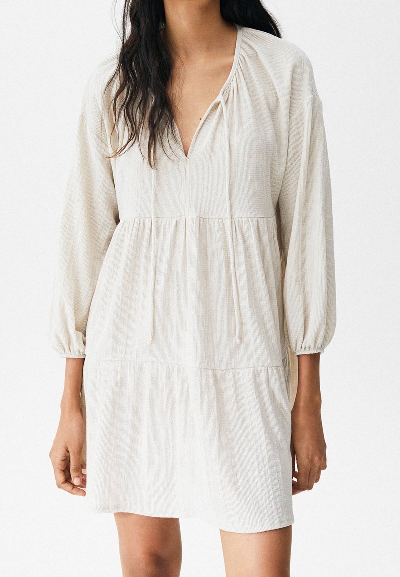 Robe ample crème avec manches longues, taille froncée et encolure en V avec liens, portée par une personne aux longs cheveux foncés.