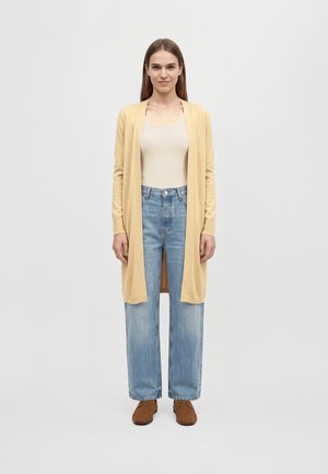 Une femme se tient face à l’objectif, sur un fond uni. Elle porte un long cardigan jaune pâle, un haut crème, un jean bleu clair et des chaussures marron.