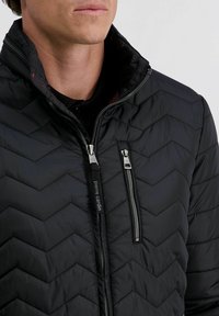 Man die een zwart gewatteerd jack draagt met twee ritsen, één verticaal op de borst en één rits in het midden met "pierre cardin" merknaam.