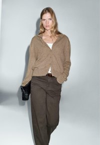 Brown cardigan with a button-down front and hood, worn over a white tank top. Paired with dark brown pants and a black clutch.
