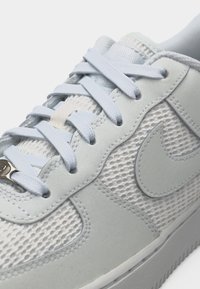 Λευκό αθλητικό παπούτσι Nike με ύφασμα από δίχτυ, σουέτ επικαλύψεις, λευκά κορδόνια και μεταλλικό τελείωμα στο κάτω κορδόνι κοντά στην περιοχή των δαχτύλων.