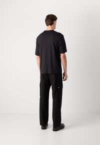 Mand står med ryggen til, iført en sort t-shirt med korte ærmer, sorte cargo-bukser med sidelommer og sorte sko på en ensfarvet baggrund.