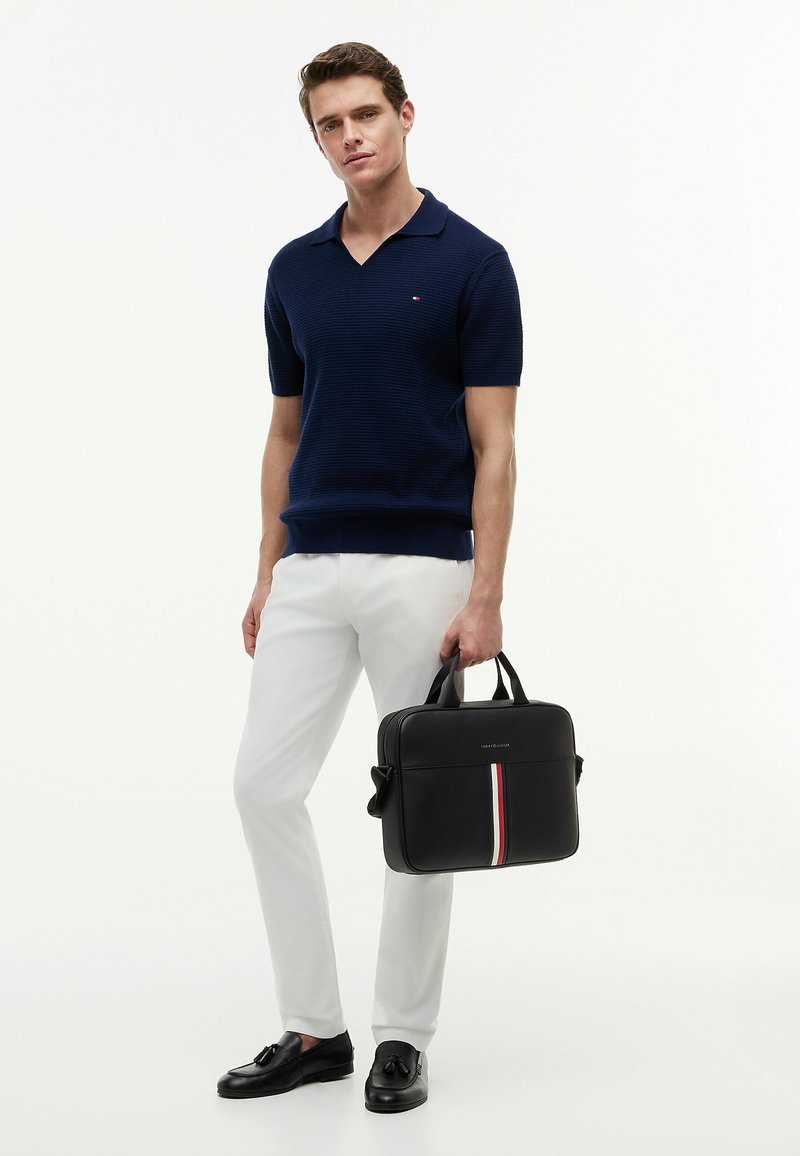 Homme en polo en maille bleu marine à manches courtes, pantalon blanc, mocassins noirs à glands, tenant une mallette noire avec une bande verticale rouge, blanche et bleue.
