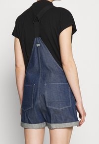 Osoba v modrých džínových šortkách s ohrnutými manžetami a černém tričku s krátkým rukávem, viděná zezadu.