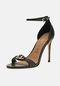 Schwarze High-Heels Sandale mit Knöchelriemen, offener Zehenpartie und goldfarbigem dekorativem Logo auf dem vorderen Riemen, beigefarbene Innensohle mit dem Markennamen GUESS.