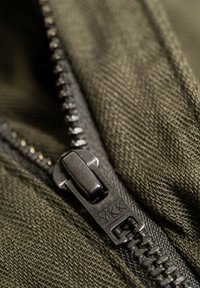 Gros plan sur un curseur de fermeture éclair YKK noir sur un tissu texturé vert olive foncé, partiellement fermé.