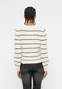 Pull crème avec des rayures horizontales grises, texture tricotée, épaules tombantes et poignets côtelés, associé à un pantalon gris foncé.