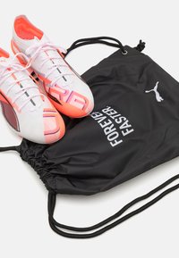 Fehér és rózsaszín foci cipők fekete részletekkel, egy fekete húzózsinóros táskával együtt, amelyen a "FOREVER FASTER" felirat fehér színben szerepel.