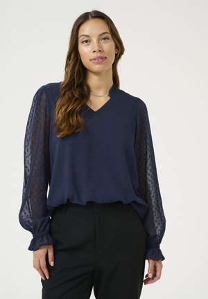 Navy blue blouse with sheer sleeves, dotted texture, and a V-neck. Features gathered cuffs and a relaxed fit. Paired with black trousers.
