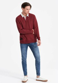 Bordeaux Sweatshirt mit Polo-Kragen und schwarzem Etikett am Saum, kombiniert mit schmal geschnittenen blauen Jeans und weißen Sneakern mit braunen Sohlen.