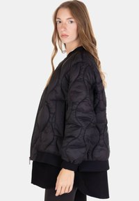 Giovane donna con lunghi capelli biondi, indossa una giacca bomber trapuntata nera e un completo nero, in piedi di profilo su uno sfondo semplice.